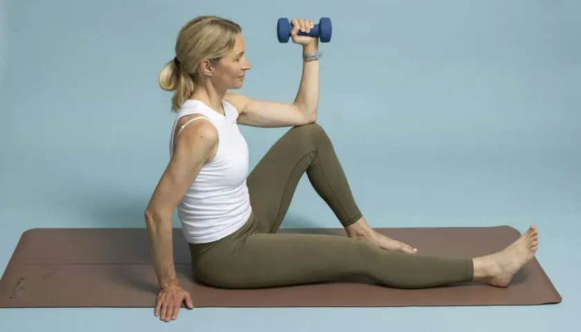 Kvinna tränar yoga nacke axlar med en hantel. Hon sitter på en brun yogamatta mot ljusblå bakgrund.