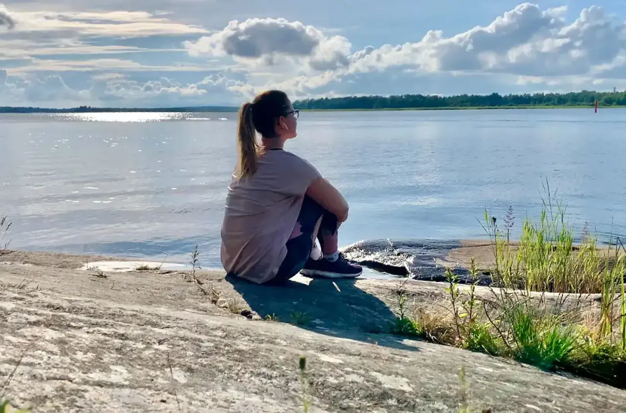 Kvinna sitter på en klippa vid havet, njuter av lugnet. Perfekt för mental avslappning och att ladda batterierna för att sova gott.