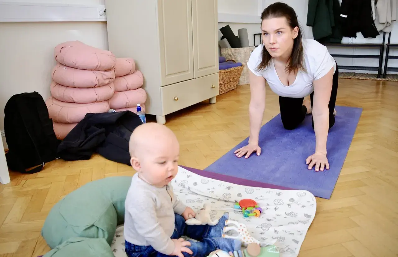 En kvinna i vit topp och svarta leggings gör baby yoga på en lila matta. En bebis sitter på en filt med leksaker.