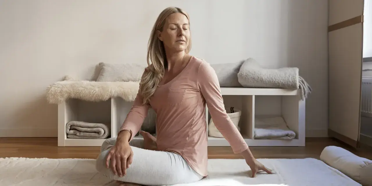 Kvinna i avslappning mindfulness-position på en matta. Bakom henne syns en hylla med filtar och kuddar.