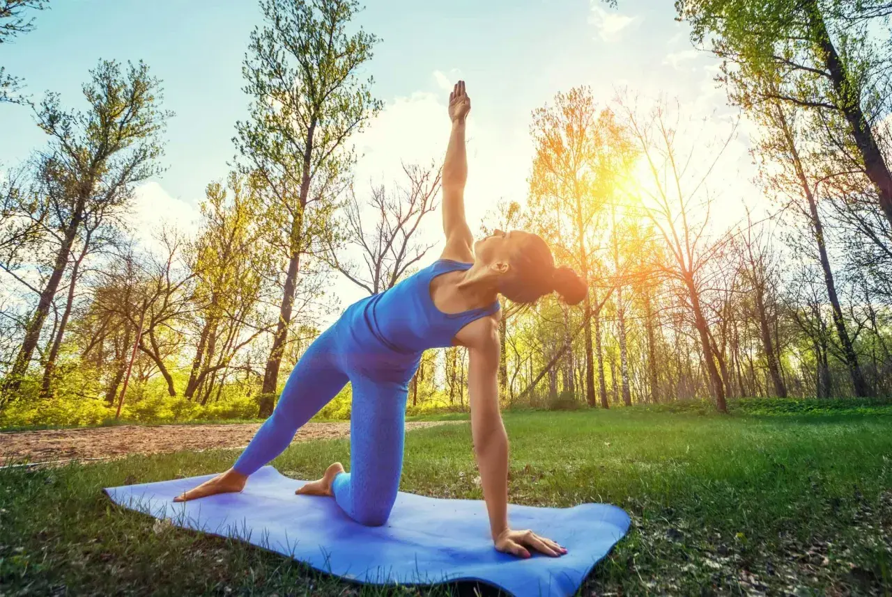 En kvinna gör yoga utomhus i en skog. Solen skiner genom träden.