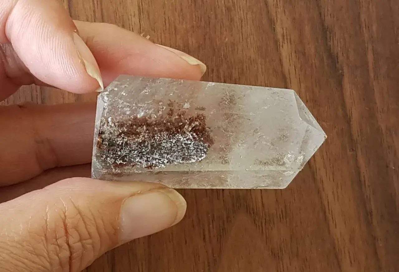 En hand håller en klar kristall med inneslutningar av rödbruna och silverfärgade mineraler, som visar lodolitens fascinerande egenskaper.