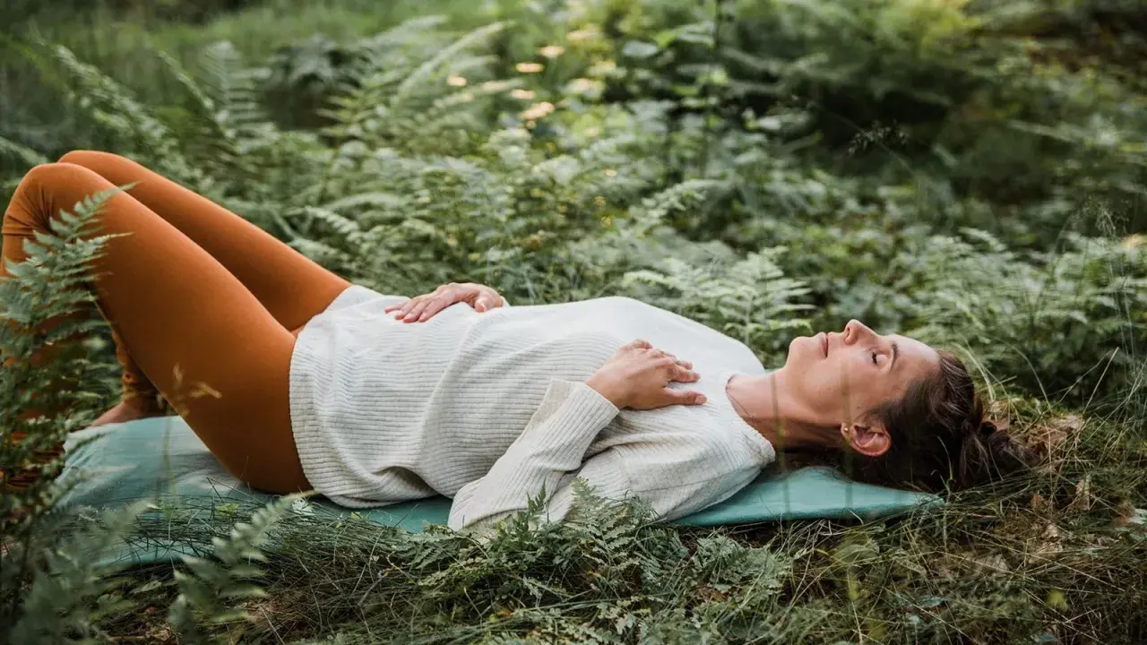 Kvinna ligger på en matta i skogen, med slutna ögon och händerna över bröstet, i en djup avslappning under yoga nidra.