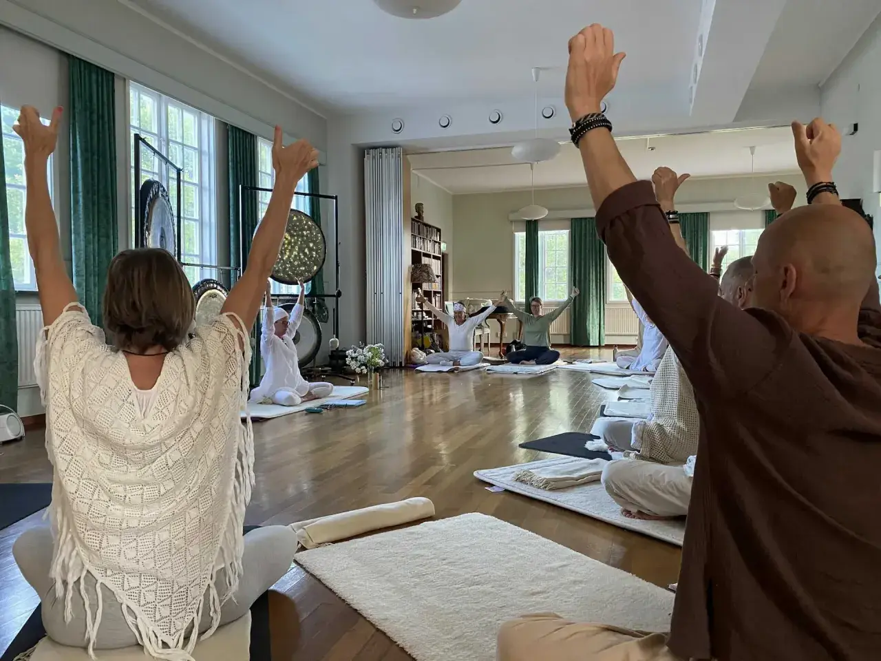 Människor sitter på mattor och höjer armarna under en kundalini yoga-klass. Gongs hänger på väggen.