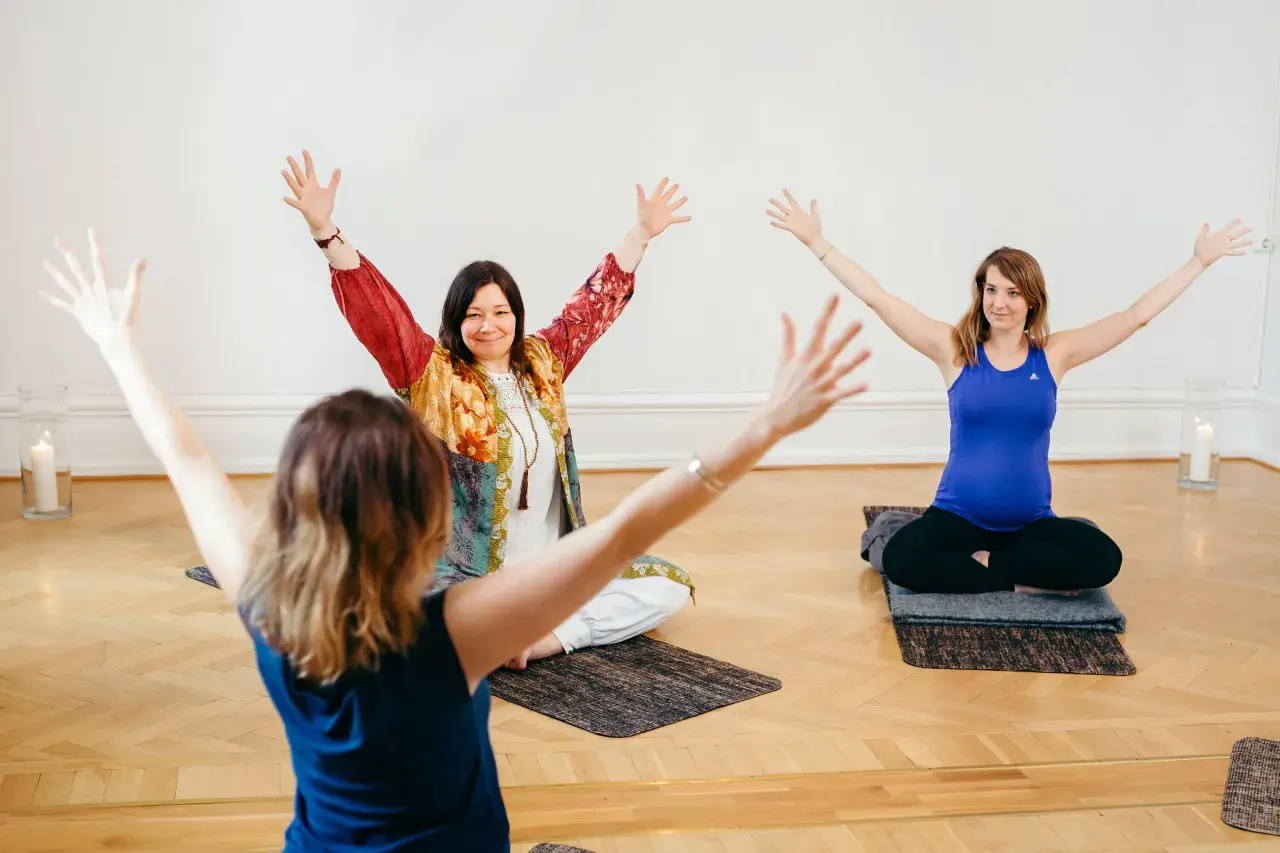 Tre kvinnor deltar i en medicinsk yoga utbildning. De sitter på mattor med armarna lyfta.