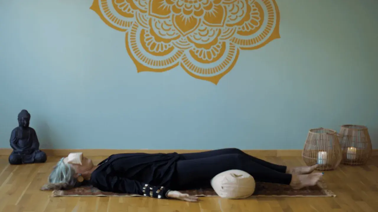 En kvinna vilar på en matta med ögonbindel, en del av en yoga nidra utbildning. En Buddha-staty och korglampor skapar en lugn atmosfär.