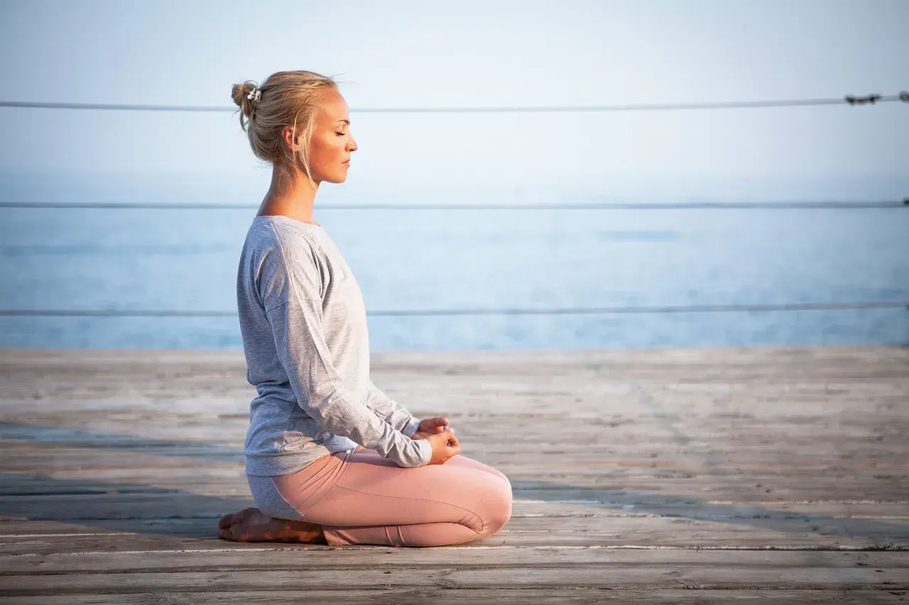 Kvinna med slutna ögon sitter i meditationsställning på en träbrygga vid havet. Hon utstrålar mental avslappning.