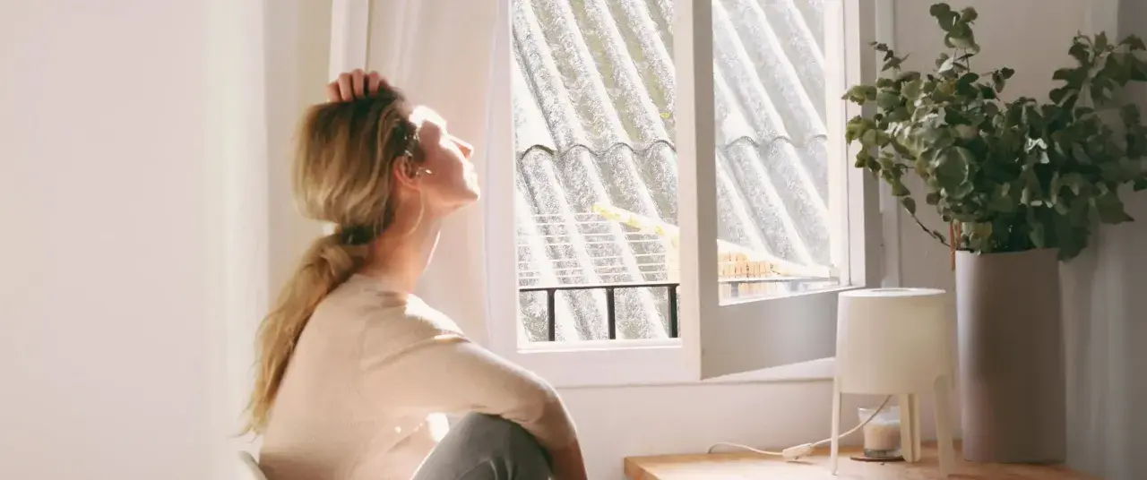 En kvinna sitter vid ett fönster, med handen i håret, och blickar uppåt. Ljuset faller på henne, men hon verkar drabbad av årstidsbunden depression.