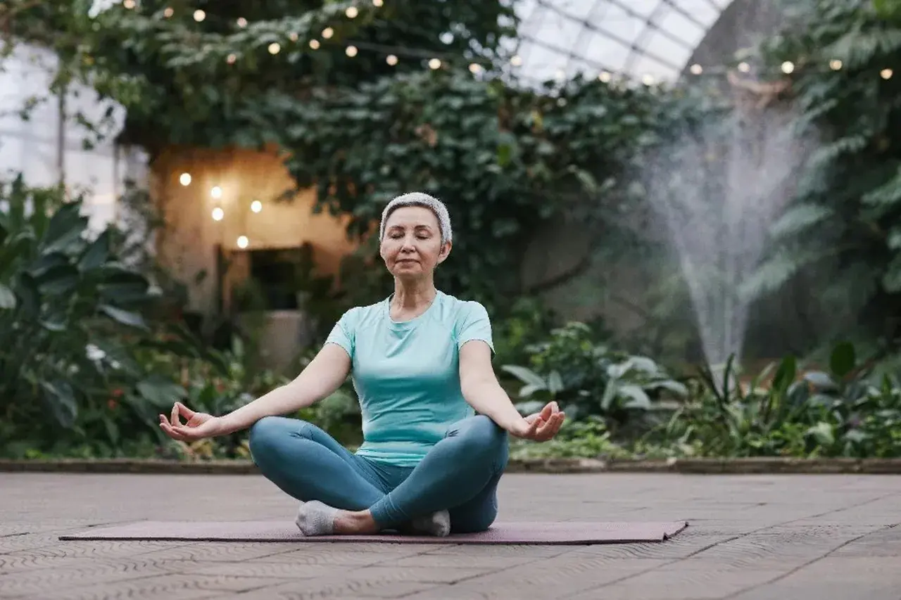 Kvinna i lotusställning, med slutna ögon och händerna i meditation, omgiven av grönska och ljusslingor. En lugn stund för meditation mot stress.