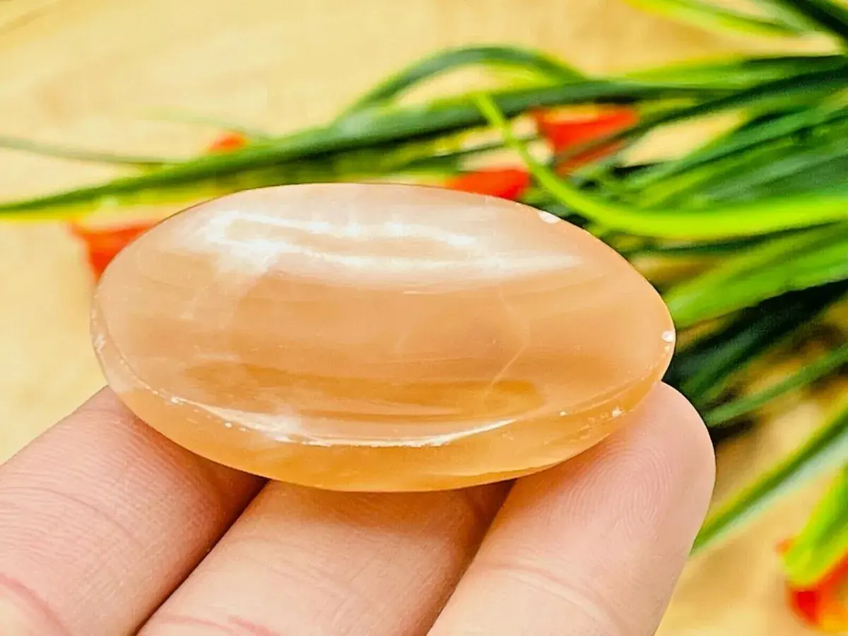 En hand håller en slät, oval honungskalcit. Dess varma, gyllene färg och mjuka lyster framhäver dess unika honungskalcit egenskaper.