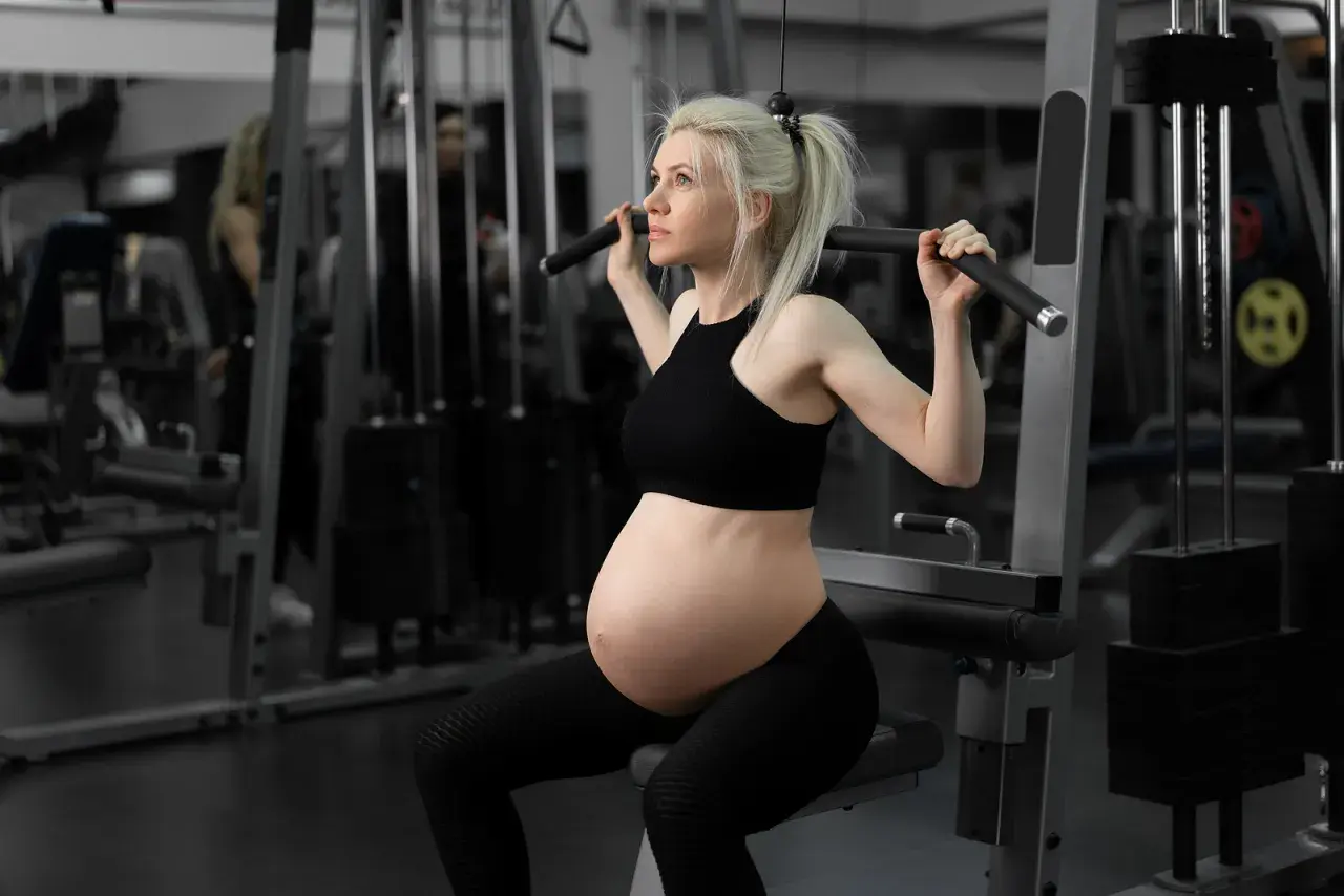 Gravid kvinna i andra trimestern tränar på gym. Hon sitter på en träningsmaskin och drar i ett handtag.