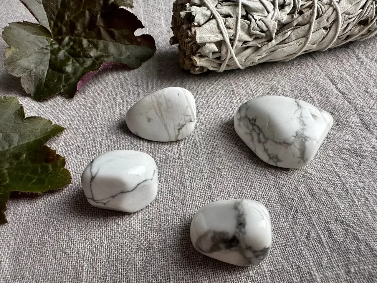 Fyra vita stenar med grå ådring, troligen magnesit, ligger på en grå duk. En bunt salvia och mörka blad ligger bredvid. Magnesit egenskaper kan vara lugnande.