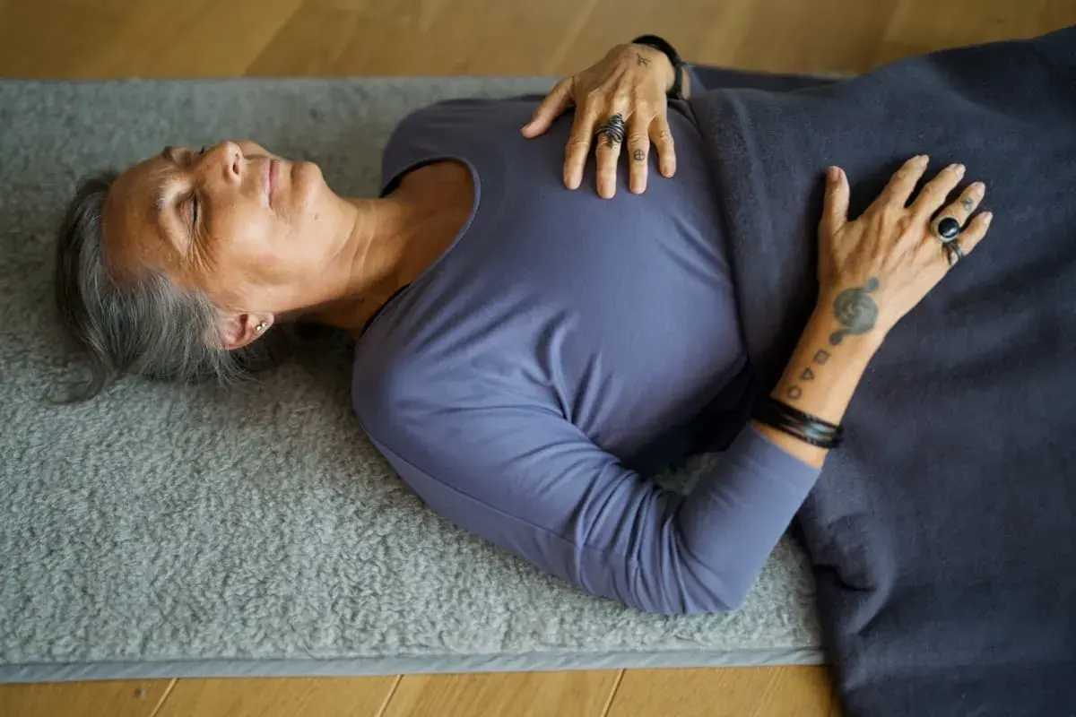 Äldre kvinna vilar på en matta, med slutna ögon och händerna över bröstet. Hon följer ett yoga nidra manus för djup avslappning.