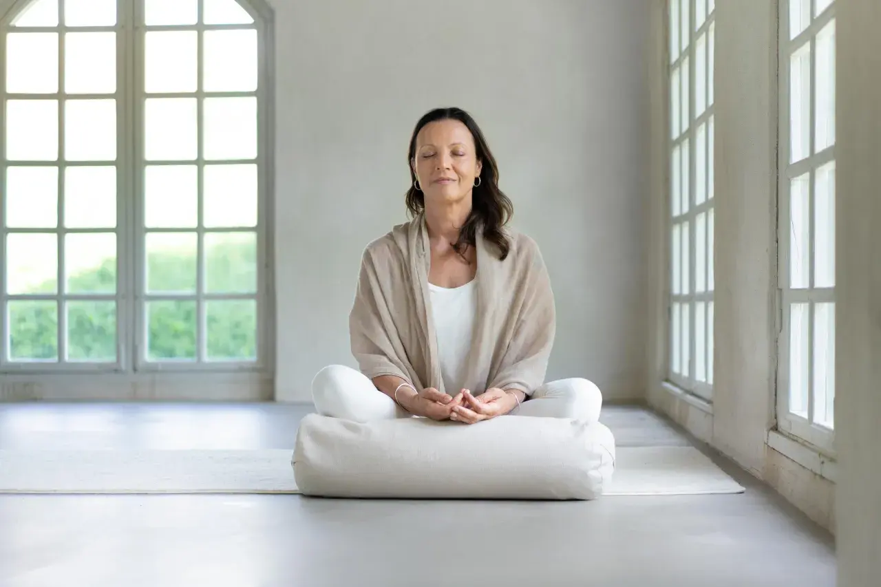 Kvinna i meditationsställning med slutna ögon, omgiven av ljus från stora fönster. Perfekt för guidad meditation text.