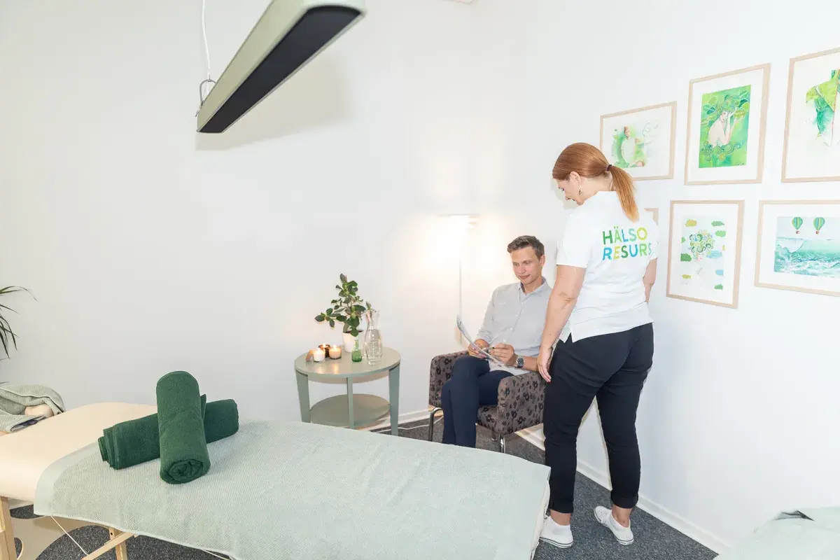 En kvinna i vit t-shirt med texten "Hälso Resurs" pratar med en man som sitter i en fåtölj. Rummet är ljust och har en massagebänk med gröna handdukar. Kanske förbereder de sig för en vagusnerven massage.