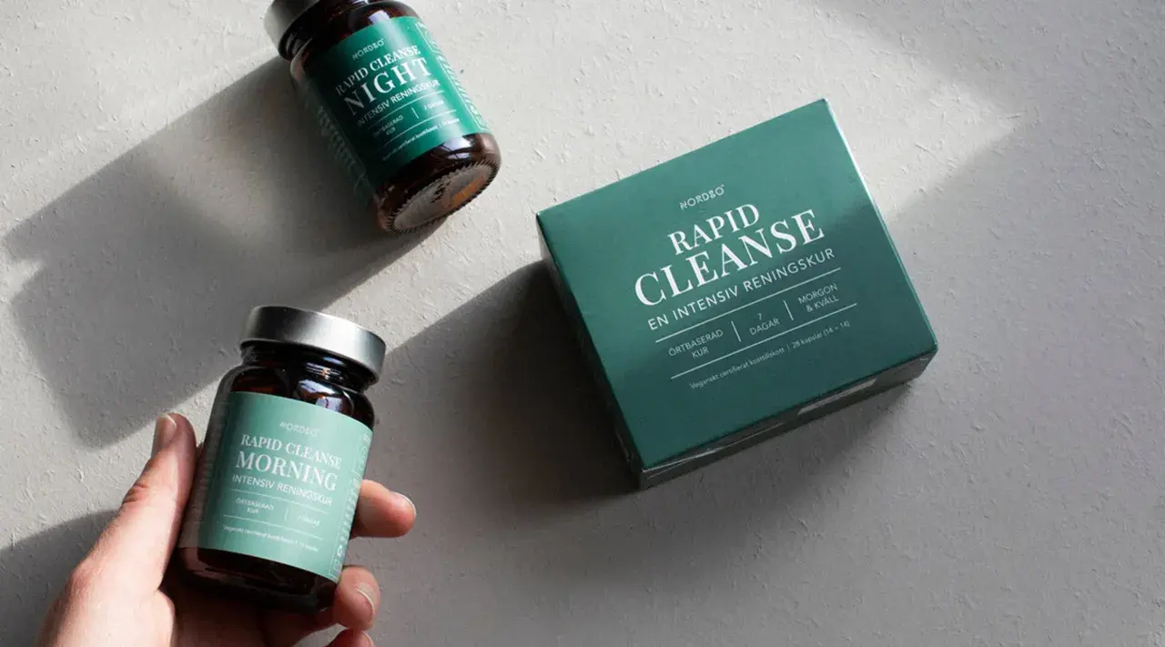 En hand håller en burk med "Rapid Cleanse Morning" kosttillskott, bredvid en låda och en burk för natten. Produkter för en intensiv rening, inspirerade av ayurvedisk medicin.