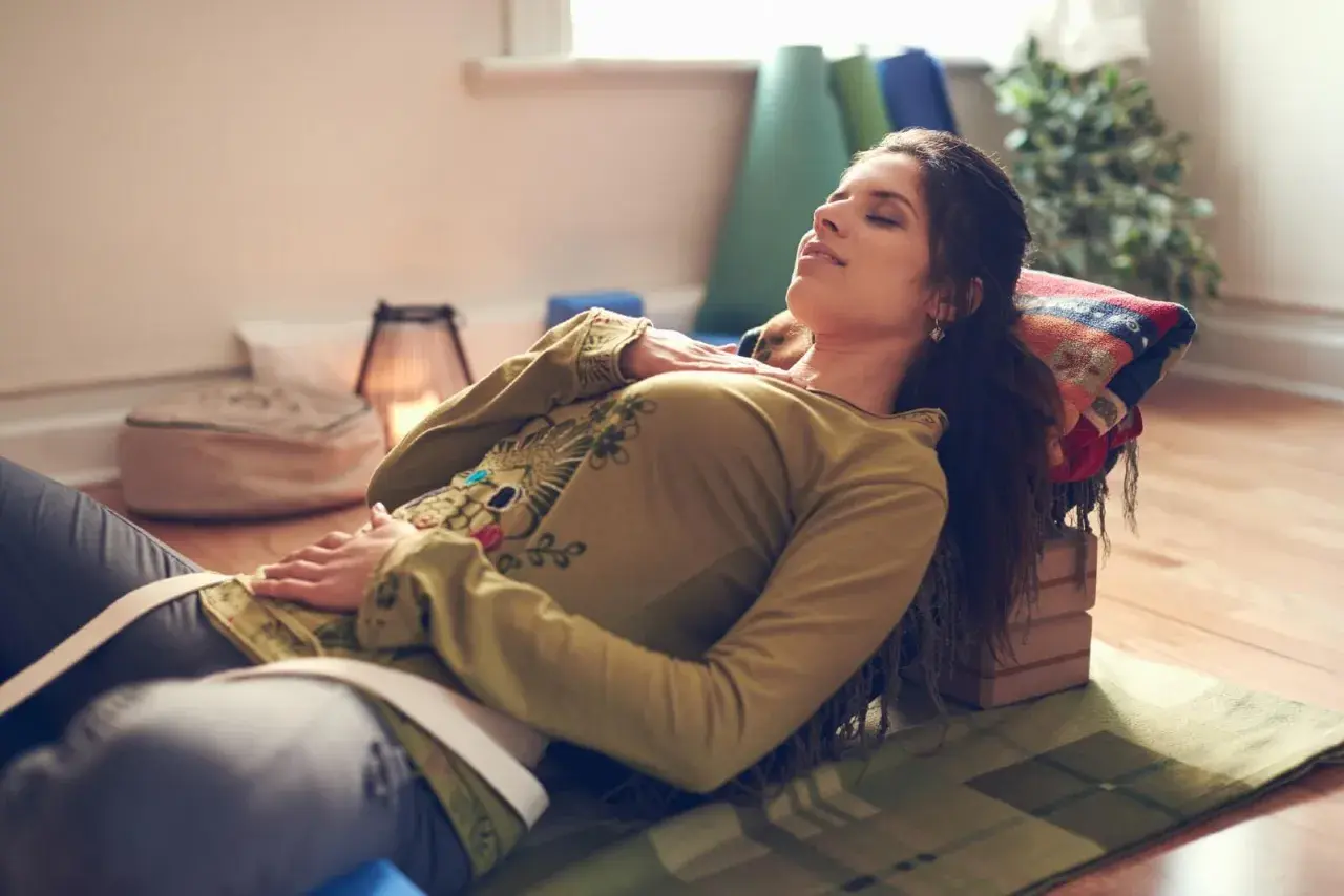Kvinna vilar med slutna ögon, upplever lugnande yoga nidra effekter. Stöd för huvud och kropp, omgiven av yogatillbehör.