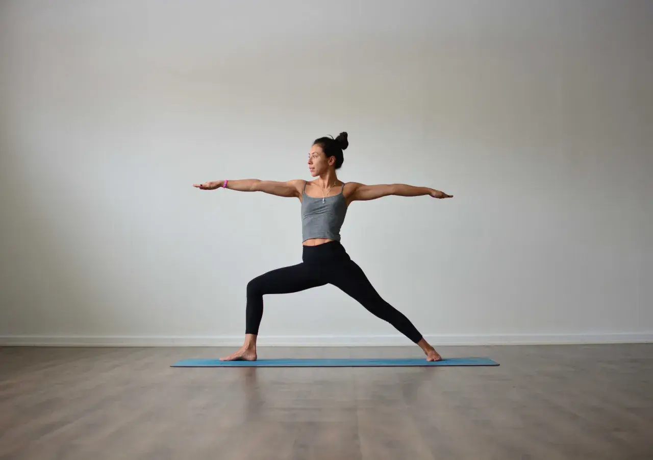 Kvinna i krigare II-ställning under vinyasa yoga. Hon står på en blå yogamatta med armarna utsträckta.