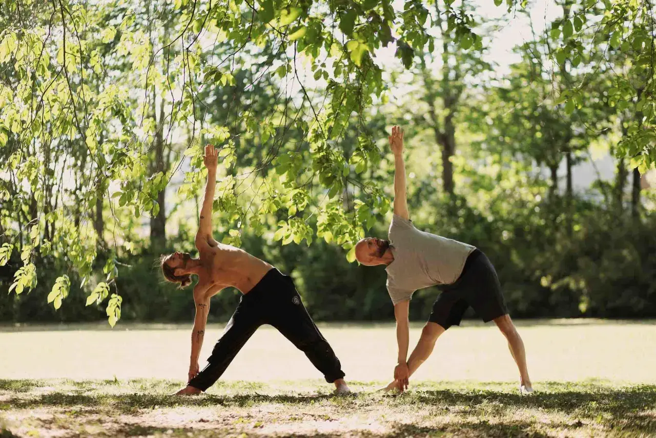 Två män utövar yoga i en park. De står i en triangelställning under trädens grönska.