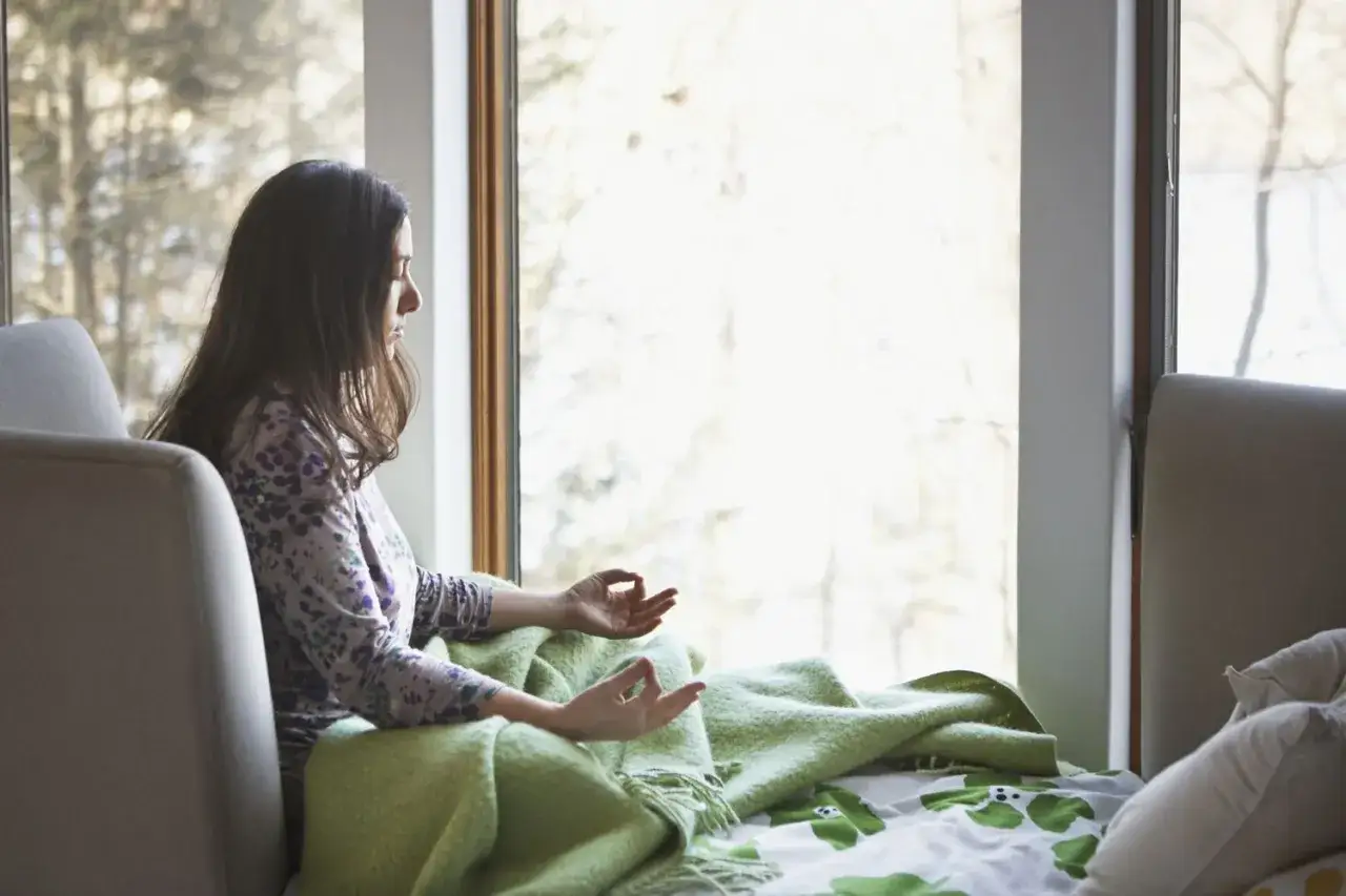 Kvinna med slutna &ouml;gon sitter i en f&aring;t&ouml;lj vid ett f&ouml;nster, insvept i en gr&ouml;n filt, och ut&ouml;var kort meditation.