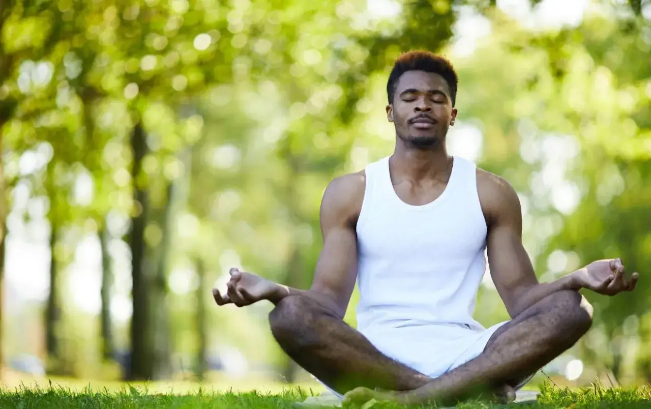 Man i vit linne sitter i lotusst&auml;llning i gr&ouml;nskande park. Han ut&ouml;var yoga och finner lugn i naturen.