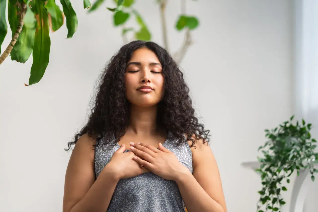 Ung kvinna med slutna &ouml;gon och h&auml;nderna &ouml;ver br&ouml;stet, en lugn stund av meditation mot stress.