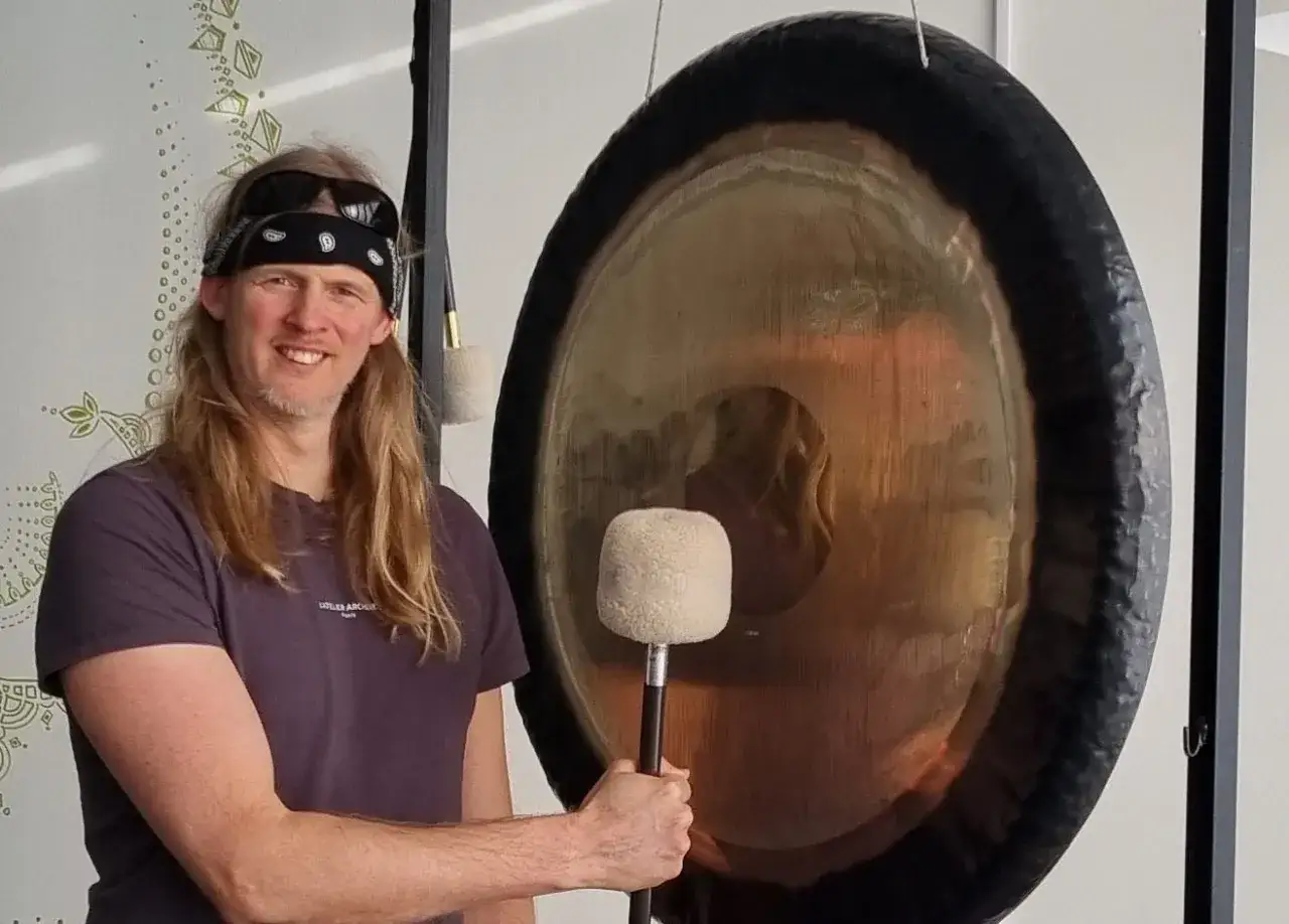 Man med långt hår och bandana håller en gong-klubba framför en stor gong. Hans leende antyder att han är mitt i en session för gongbad forskning.