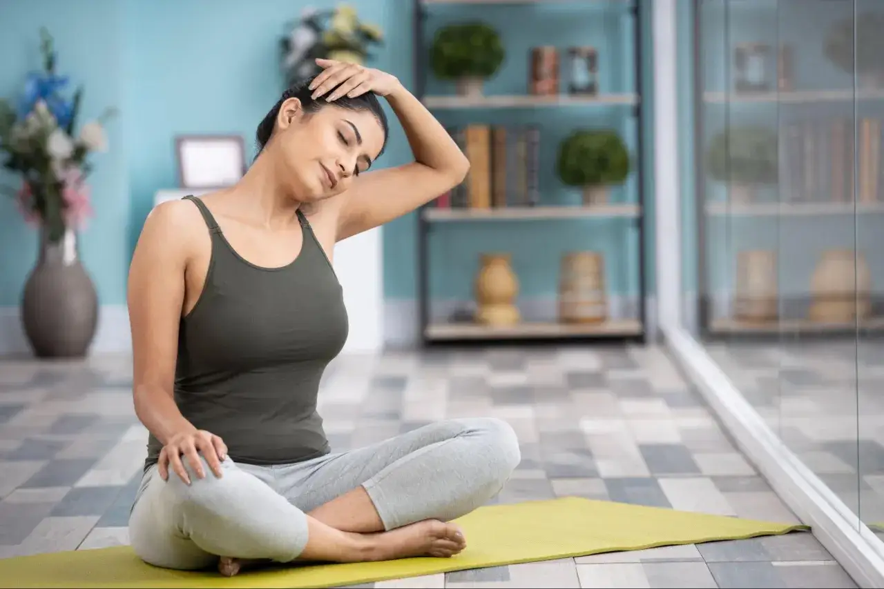 En kvinna sitter p&aring; en yogamatta och str&auml;cker f&ouml;rsiktigt sin nacke f&ouml;r att slappna av i nacken.