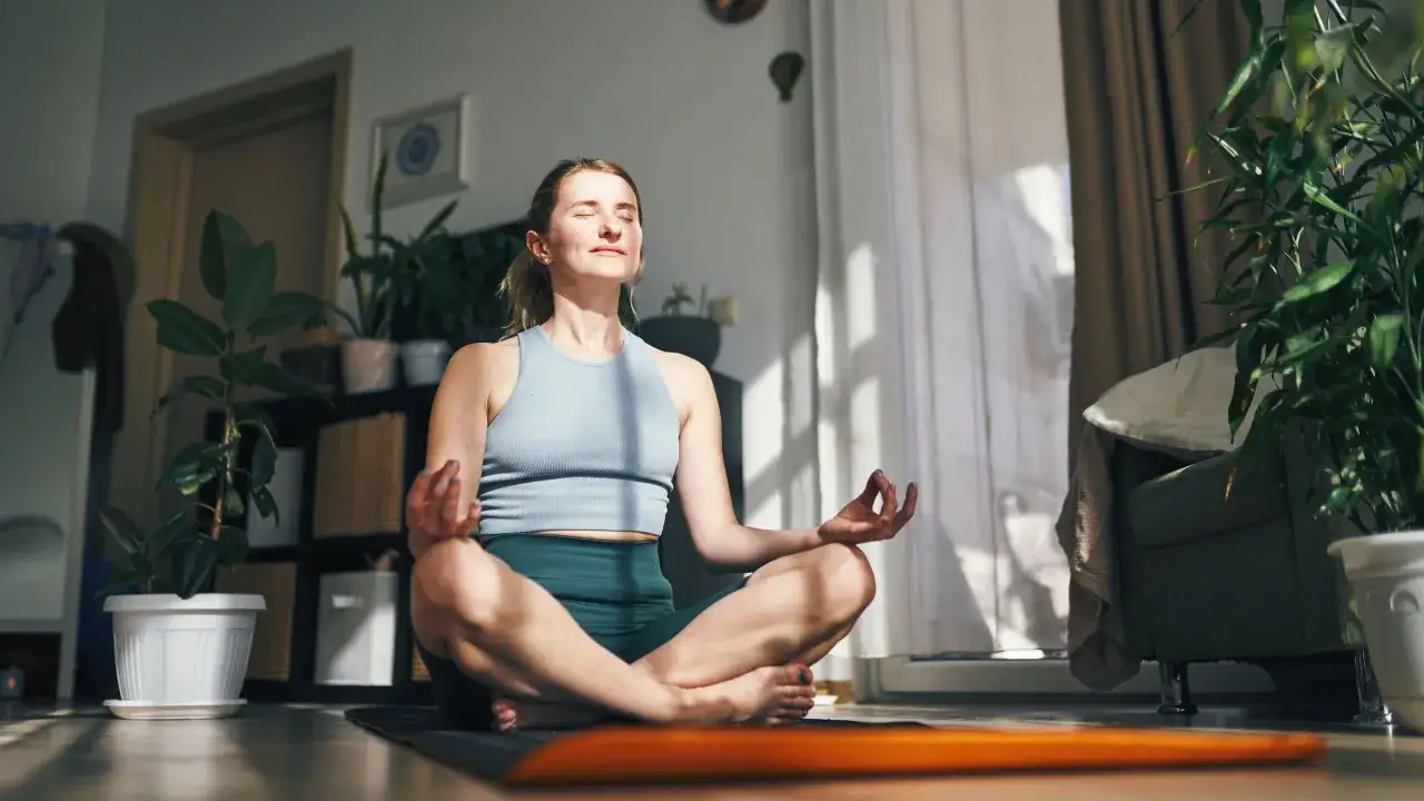 Kvinna med slutna &ouml;gon sitter i lotusst&auml;llning p&aring; en yogamatta. Solstr&aring;lar lyser in i rummet. En stund av mindfulness i vardagen.