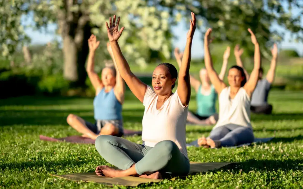 En grupp m&auml;nniskor ut&ouml;var yoga utomhus. En kvinna i f&ouml;rgrunden sitter i meditationsst&auml;llning med armarna lyfta, en &ouml;vning som kan vara en del av ayurvedisk medicin.