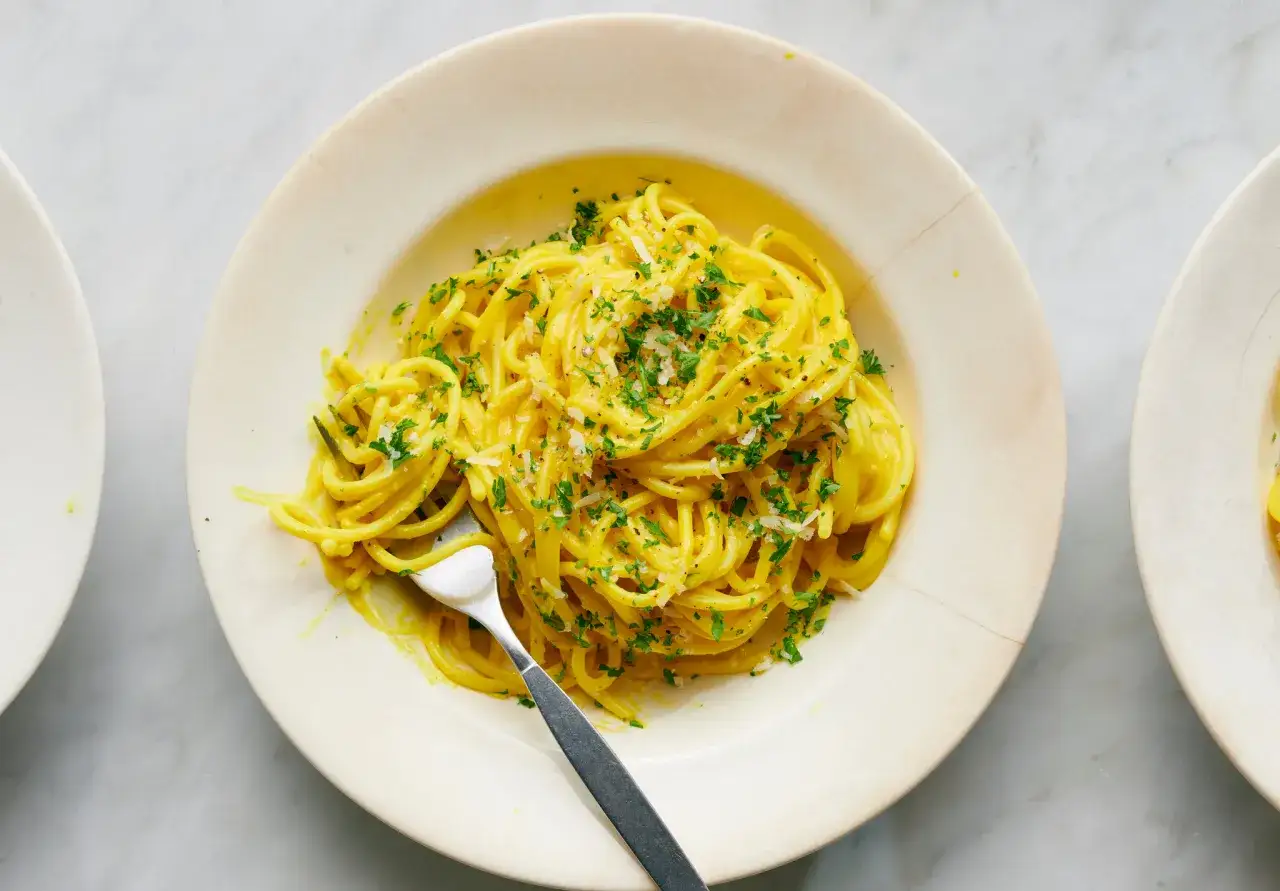 En tallrik med gyllene spaghetti, en h&auml;rlig gurkmeja pasta, toppad med f&auml;rsk persilja och riven ost. En gaffel ligger redo att avnjutas.