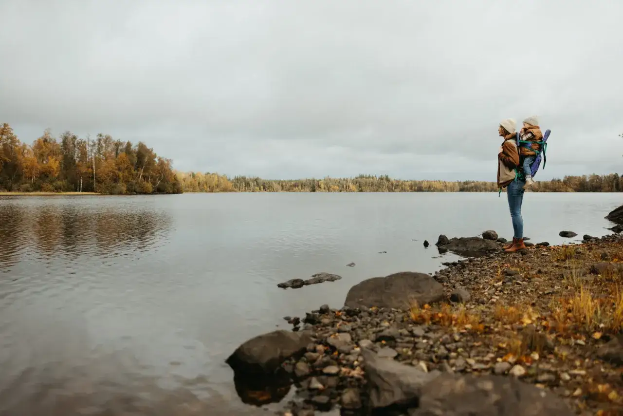 En person b&auml;r ett barn i b&auml;rsele vid en sj&ouml; i Sm&aring;land. H&ouml;stf&auml;rger syns i skogen bakom. En bl&aring; zon av lugn.