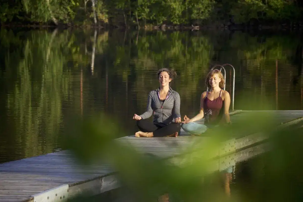 Tv&aring; kvinnor mediterar p&aring; en brygga vid en spegelblank sj&ouml;. En perfekt yoga helg i naturen.
