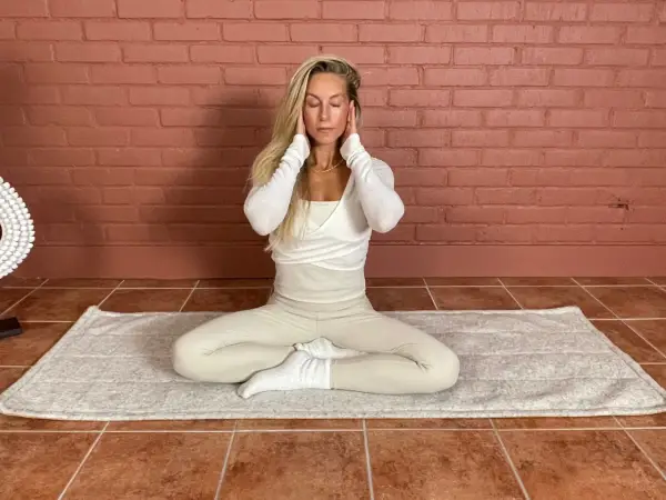 Kvinna med slutna ögon sitter i meditationsställning på en matta. Hon har händerna vid tinningarna. Detta kan vara en del av en lymfyoga utbildning.